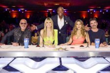 Five judges sit at a panel table on a live TV talent show, smiling at the camera as the host stands behind them with open arms.