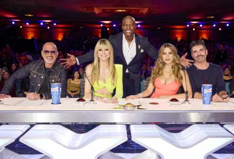 Five judges sit at a panel table on a live TV talent show, smiling at the camera as the host stands behind them with open arms.