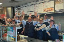 Group of restaurant staff behind the counter waving at the camera, wearing aprons and uniforms.
