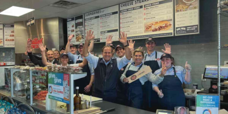 Group of restaurant staff behind the counter waving at the camera, wearing aprons and uniforms.