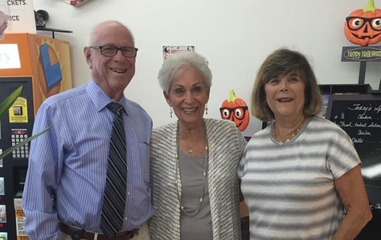 Three seniors pose indoors with Halloween decorations behind them; left: man in a blue striped shirt and tie, center: woman in a light cardigan, right: woman in a striped gray top.