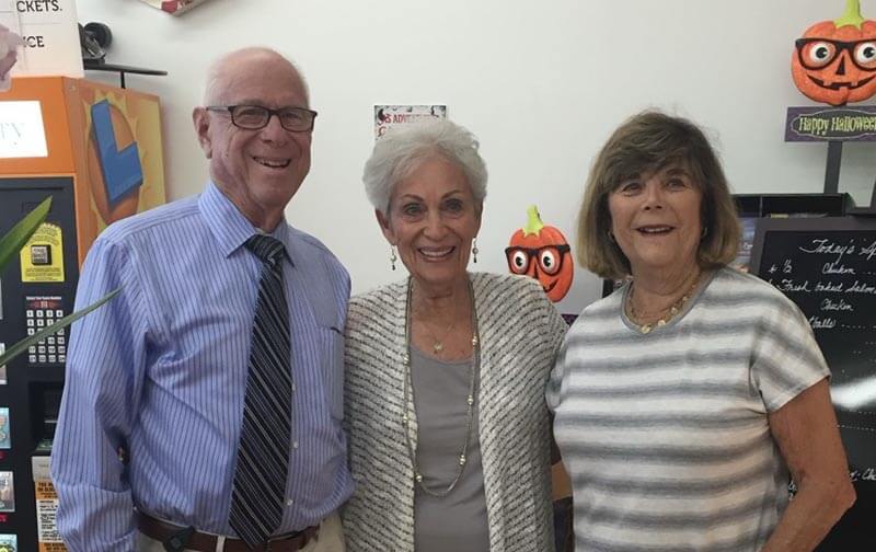 Three seniors pose indoors with Halloween decorations behind them; left: man in a blue striped shirt and tie, center: woman in a light cardigan, right: woman in a striped gray top.