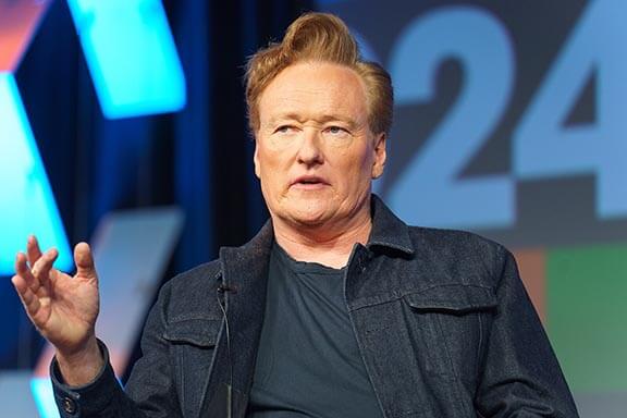 Conan O'Brien in a denim jacket speaking on stage, gesturing with his hand; large blue lights and a '24' in the background.