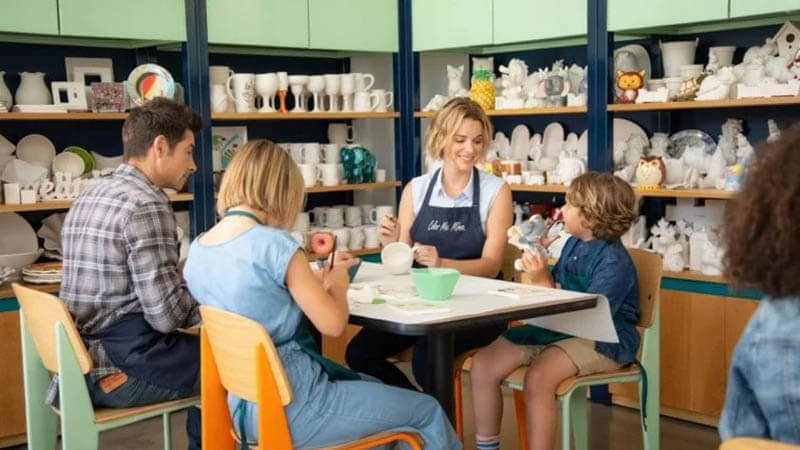 Instructor at Color Me Mine guides a pottery-painting class with kids and adults around a table with ceramic pieces in the background.
