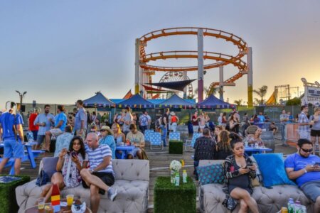 Off The Hook Santa Monica Seafood Festival | Santa Monica Sun Crowd at Santa Monica Pier attends an outdoor festival on a wooden deck, with blue seating under tents near roller-coaster track at sunset.