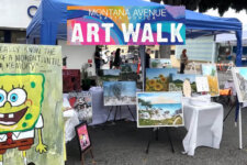 Outdoor art market booth with a colorful 'Art Walk' banner, displaying framed landscape paintings and a SpongeBob cutout on the left.