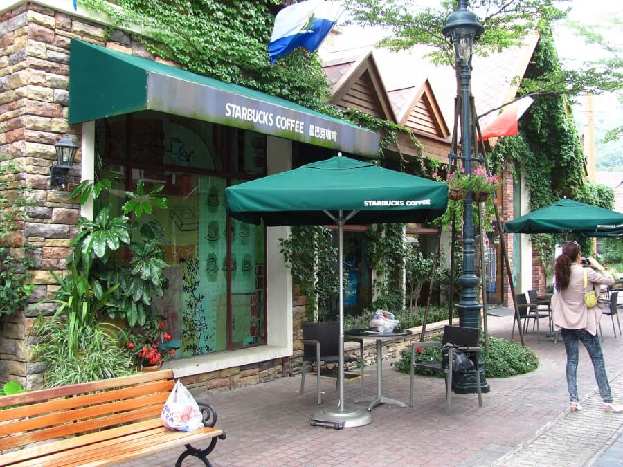 Starbucks Coffee storefront with green awning, outdoor seating under umbrellas, stone wall, and lush plants; a woman takes a photo nearby.