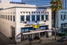Two-story beige urban building with California Pizza Kitchen storefront and glass doors on a sunny street, palm tree nearby.