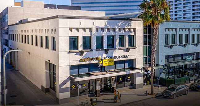 Two-story beige urban building with California Pizza Kitchen storefront and glass doors on a sunny street, palm tree nearby.