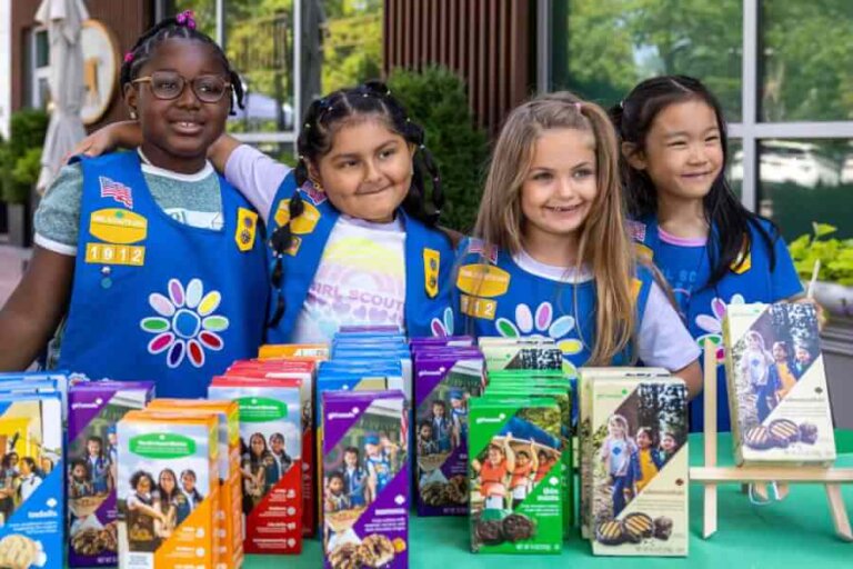 Four diverse young girls in blue scout sashes stand behind a table full of colorful boxed cookies