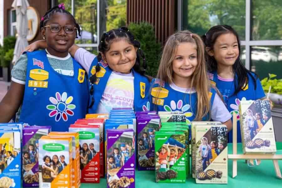 Four diverse young girls in blue scout sashes stand behind a table full of colorful boxed cookies
