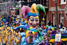 Colorful parade float featuring a large smiling jester wearing a multicolored hat, with spectators lining the street beyond.