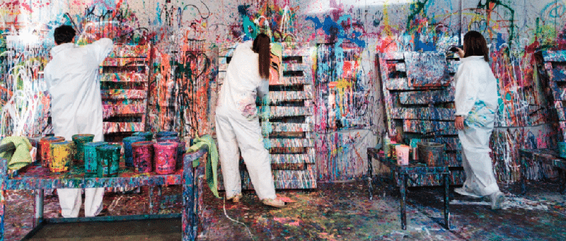 Three artists in white coveralls paint a wall splattered with bright colors in a messy art studio, with paint cans on tables nearby.