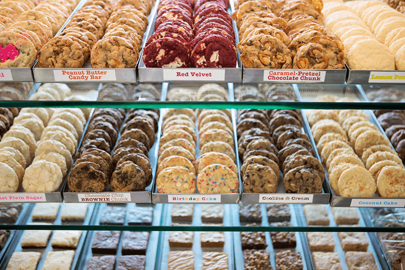 Cookie Good display | Santa Monica Sun Bakery display case filled with rows of assorted cookies. Top row includes Peanut Butter Candy Bar, Red Velvet, and Caramel-Pretzel. Bottom row features Chocolate Chip Brownie, Birthday Cake, and Cookies & Cream.