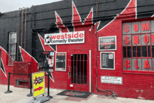 Front of Westside Comedy Theater with a bold black-and-red mural, red brick, and the entrance door guarded by bars; signage and a sidewalk A-frame directing to the venue.