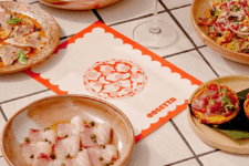Assorted tapas-style dishes surround a central scalloped red menu card on a tiled table, including sliced meat with capers, a grain salad, and bruschetta.