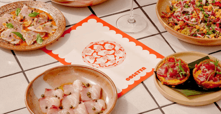 Assorted tapas-style dishes surround a central scalloped red menu card on a tiled table, including sliced meat with capers, a grain salad, and bruschetta.