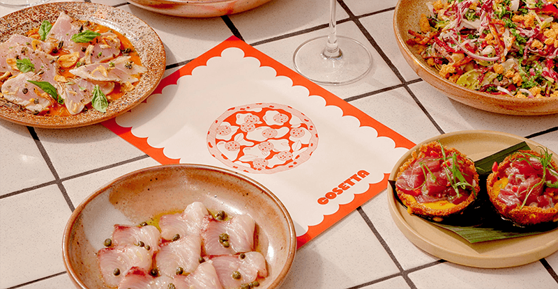 Assorted tapas-style dishes surround a central scalloped red menu card on a tiled table, including sliced meat with capers, a grain salad, and bruschetta.