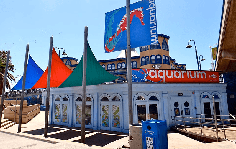 Exterior view of an aquarium building with white arched windows, bold orange sign, and colorful sail shade structures on a sunny day.
