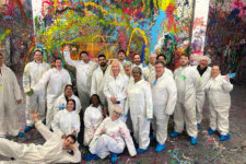 Splatter Studio LA customers | Santa Monica Sun Diverse group of people in white coveralls pose for a photo in a vibrant, paint-splattered art studio.