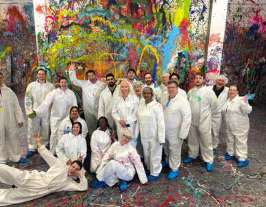 Diverse group of people in white coveralls pose for a photo in a vibrant, paint-splattered art studio.