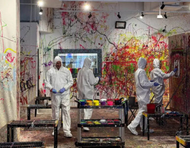 People in white protective suits and gloves painting a wall covered in colorful splatters in a paint-splatter art studio.