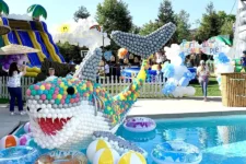 Balloon Celebrations pool | Santa Monica Sun Colorful balloon shark sculpture in a pool at a sunny outdoor carnival, with slides and people in the background.