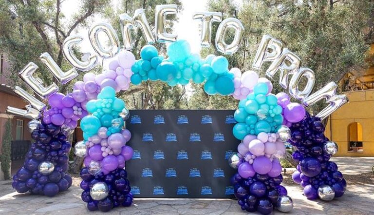 Balloon arch spelling 'Welcome to Prom' over a blue backdrop for a high school prom event.