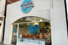 Storefront for 'cookie good' with a blue circular sign, white brick exterior, and pumpkins on a counter inside the curved window opening