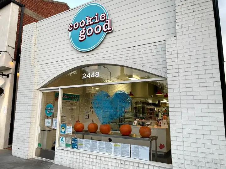 Storefront for 'cookie good' with a blue circular sign, white brick exterior, and pumpkins on a counter inside the curved window opening