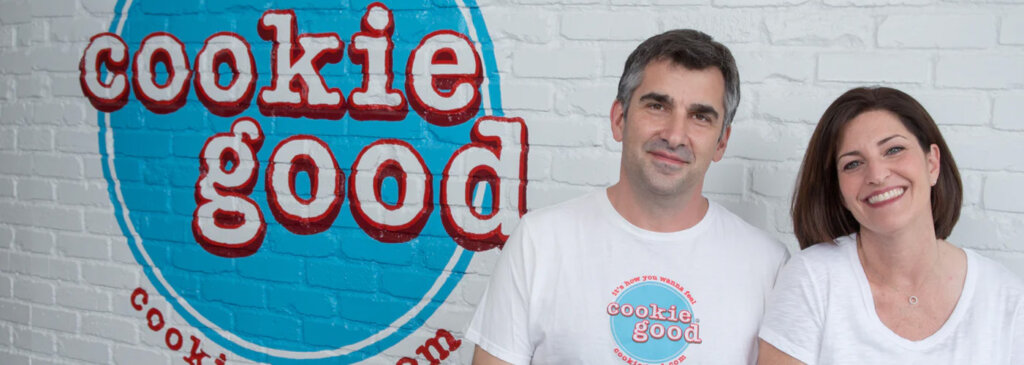 Two adults smile at the camera in white shirts with a Cookie Good logo, standing in front of a white brick wall with a large blue circular logo behind them.