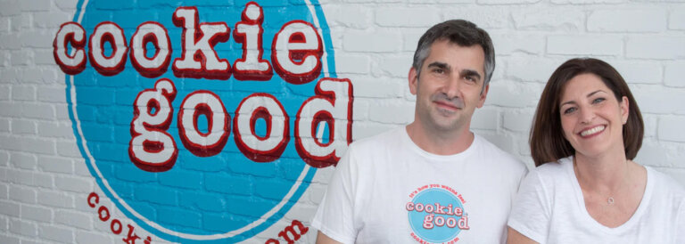 Two adults smile at the camera in white shirts with a Cookie Good logo, standing in front of a white brick wall with a large blue circular logo behind them.