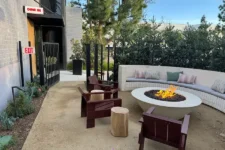 Outdoor lounge area with a round fire pit, curved bench seating, and wooden chairs around a lit fire pit.