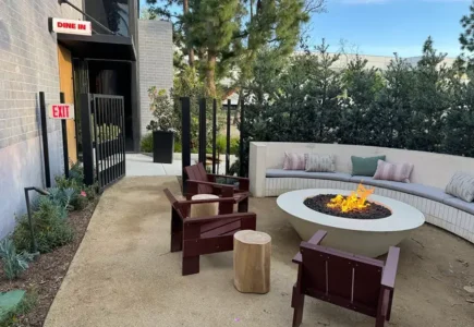 Outdoor lounge area with a round fire pit, curved bench seating, and wooden chairs around a lit fire pit.