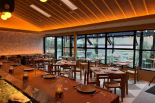 Bright, modern restaurant with wood tables set for diners, bar counter in foreground, and large windows overlooking an outdoor patio at dusk/warmer lighting.