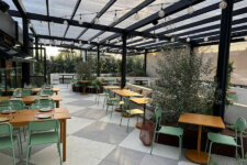 Open-air dining patio with wooden tables, mint-green chairs, and string lights under a transparent roof.