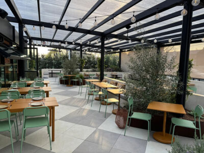 Open-air dining patio with wooden tables, mint-green chairs, and string lights under a transparent roof.