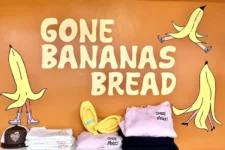 Colorful storefront display with the sign 'Gone Bananas Bread' and cartoon bananas on an orange wall, plus shelves of clothes and accessories below.