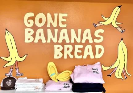 Colorful storefront display with the sign 'Gone Bananas Bread' and cartoon bananas on an orange wall, plus shelves of clothes and accessories below.