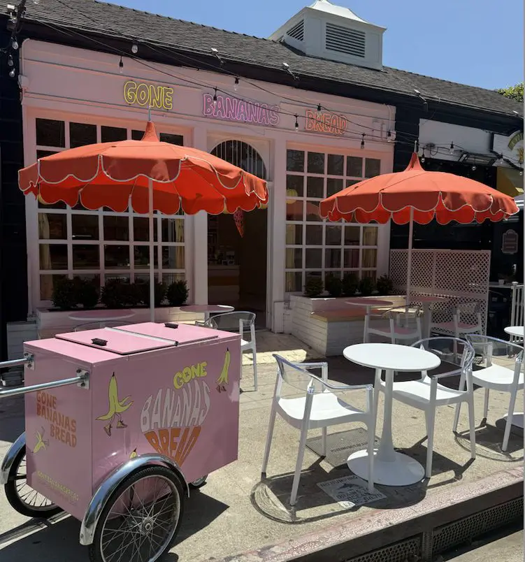 Pink bakery storefront 'Gone Bananas Bread' with white-framed windows, string lights, and outdoor seating under orange umbrellas.