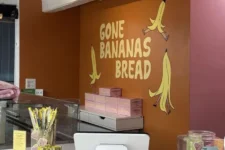 Bakery counter with orange wall reading 'Gone Bananas Bread' and banana illustrations, pink boxes on the shelf, and a white point-of-sale system on the counter