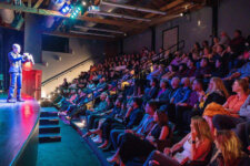 Illusion Magic Lounge audience | Santa Monica Sun Speaker in a suit delivers a talk at a microphone on a lit stage, facing a packed theater audience of adults and teens.