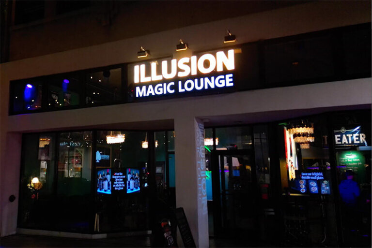 Night view of Illusion Magic Lounge storefront with illuminated sign over glass doors and windows showing interior lighting and decor at street level at night.