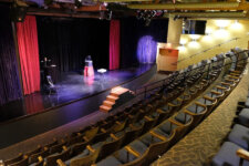 Illusion Magic Lounge theater | Santa Monica Sun Empty theater with a small stage, red and black curtains, a lectern, and a few chairs; empty audience seating surrounds the stage.