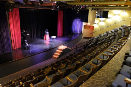 Empty theater with a small stage, red and black curtains, a lectern, and a few chairs; empty audience seating surrounds the stage.