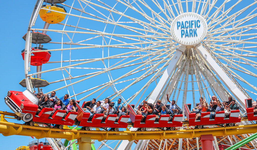 Red roller-coaster train carrying riders on a track beneath a large Ferris wheel at Pacific Park, with a clear blue sky above.