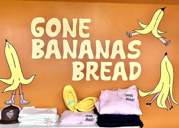 Bright orange display wall with the bold words 'GONE BANANAS BREAD' and banana illustrations; shelves hold folded clothes and yellow sandals.