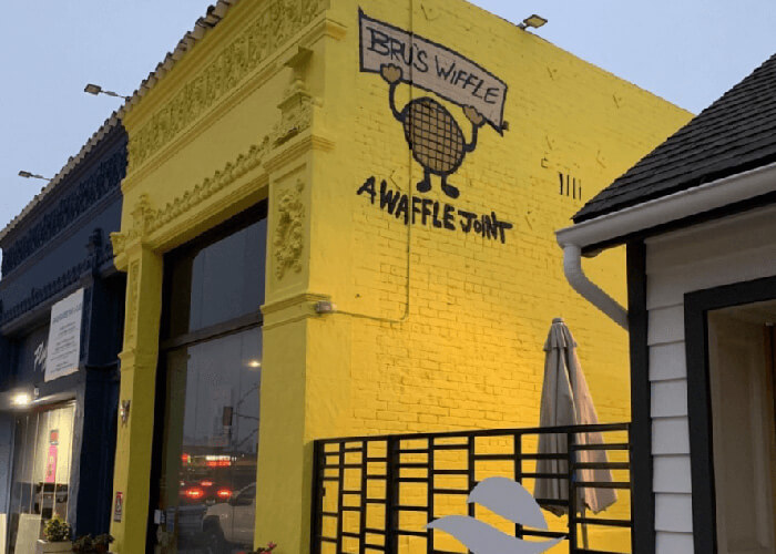 Yellow brick storefront with a mural featuring a cartoon waffle holding a banner above the text 'A WAFFLE JOINT'; outdoor seating railing and a neighboring blue-painted building are visible.