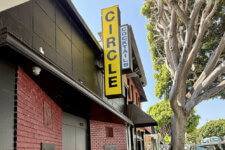 Yellow vertical 'CIRCLE' sign on a brick storefront with a blue 'COCKTAILS' sign and a tree-lined sidewalk on the right.