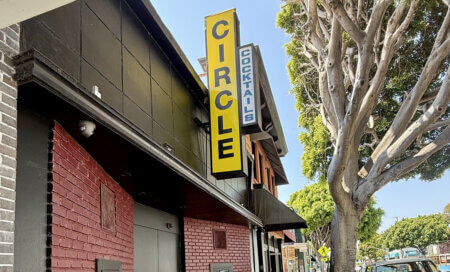 Yellow vertical 'CIRCLE' sign on a brick storefront with a blue 'COCKTAILS' sign and a tree-lined sidewalk on the right.